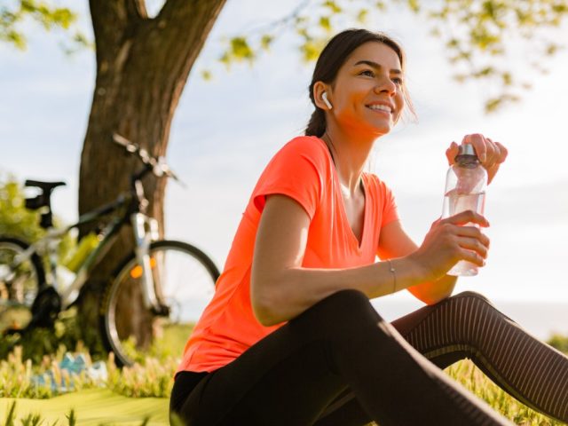 smiling beautiful woman drinking water in bottle doing sports in morning in park nature doing yoga on mat, pink fitness outfit, happy healthy lifestyle, music in earphones, bicycle on background